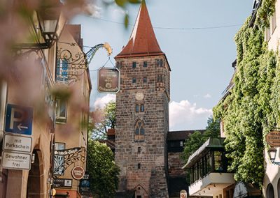 Blick auf einen Turm durch eine mit Schildern gesäumte Straße, Café-Terrassen im Vordergrund.