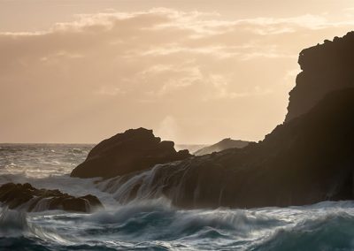 Felsige Küste im Gegenlicht mit bewegtem Meer und warmer Abendstimmung.