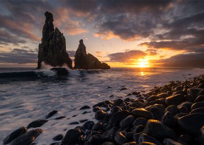 Felsformationen im Meer bei Sonnenuntergang mit dramatischem Himmel und Wellen an einer steinigen Küste.