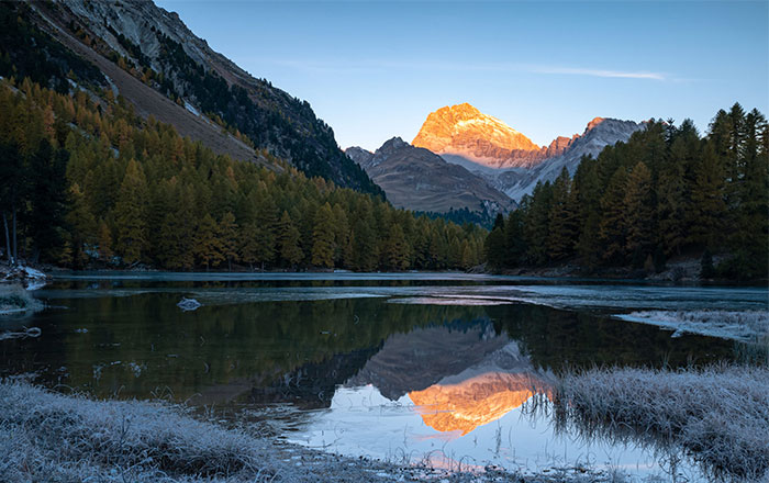 Berglandschaft mit spiegelndem See und beleuchtetem Gipfel im Morgenlicht.