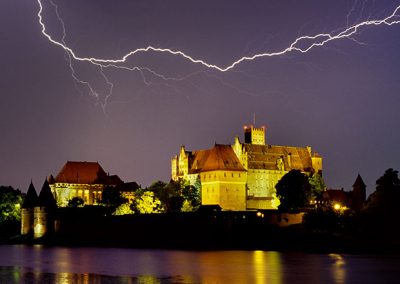 Beleuchtete Burg bei Nacht mit Blitzen am Himmel und Spiegelung im Wasser.
