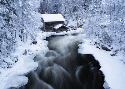 Holzhaus an einem fließenden Fluss, umgeben von verschneiten Bäumen.