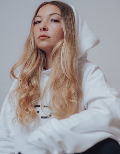 Junge Frau mit blondem Haar in weißem Hoodie, schaut selbstbewusst nach vorne.