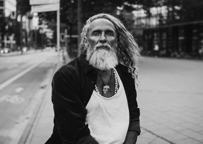Älterer Mann mit weißem Bart und langen Haaren steht auf einer leeren Straße.