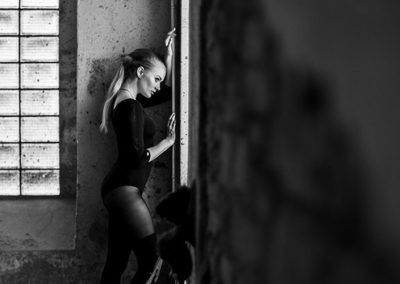 Frau lehnt in einem Bodysuit an einer Wand mit Glasbausteinen, weiches Licht, monochrom.