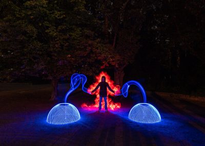 Person mit ausgebreiteten Armen, umgeben von roten Flammen und blauen Lichtbögen im Dunkeln.