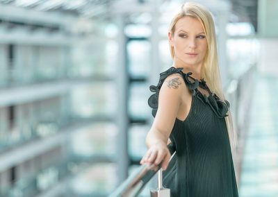 Blonde Frau in dunklem Kleid mit Tattoo posiert an Glasgeländer in heller, moderner Architektur