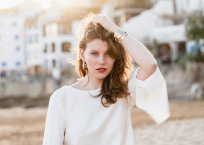 Frau im weißen Outfit steht im weichen Sonnenlicht und berührt ihr Haar, Strandkulisse im Hintergrund.