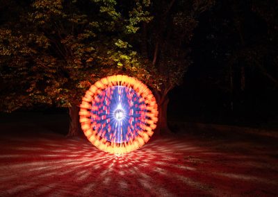 Kreis aus leuchtendem Licht in Blau, Orange und Weiß vor Bäumen bei Nacht.