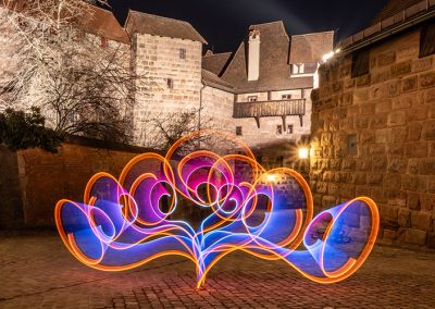 Verschnörkelte Lichtform in Orange, Blau und Violett vor Nürnberger Altstadtmauer.