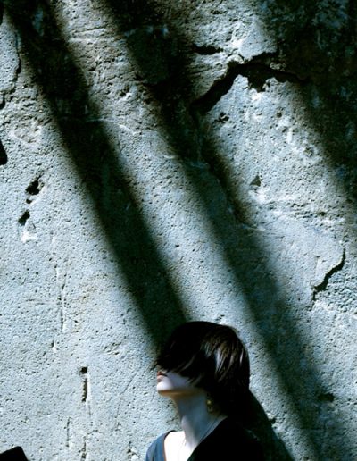 Frau mit kurzen Haaren sitzt im Schattenwurf vor grober Betonwand.
