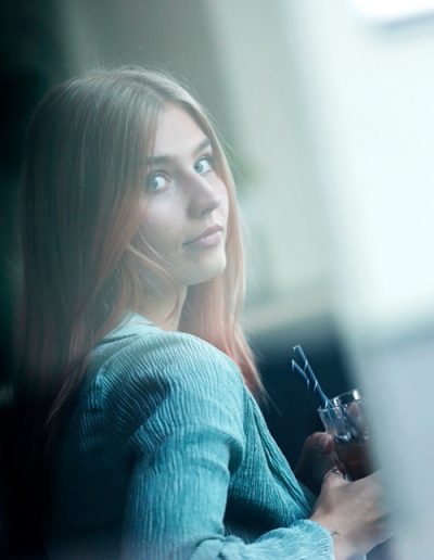 Junge Frau mit roséfarbenem Haar blickt über die Schulter durch ein Fenster, Glas spiegelt Licht.