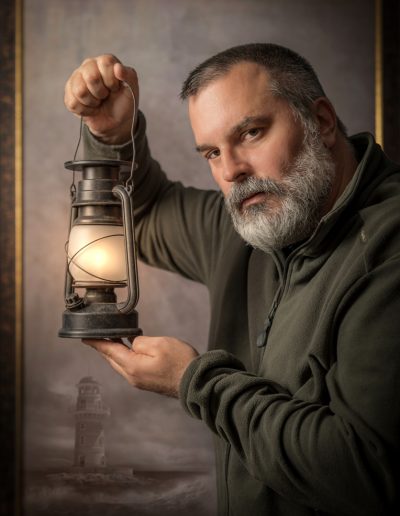 Mann mit grauem Bart hält eine alte Laterne mit leuchtender Flamme, gemalter Leuchtturm im Hintergrund.