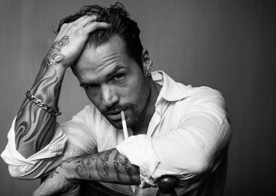 Mann mit Tattoos, Zigarette im Mund, Blick in die Kamera, rebellischer Ausdruck.