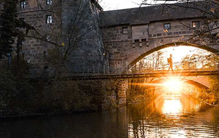 Ein Spaziergänger auf einer historischen Brücke in Nürnberg, während die untergehende Sonne golden durch das Mauerwerk scheint und sich im Wasser spiegelt.