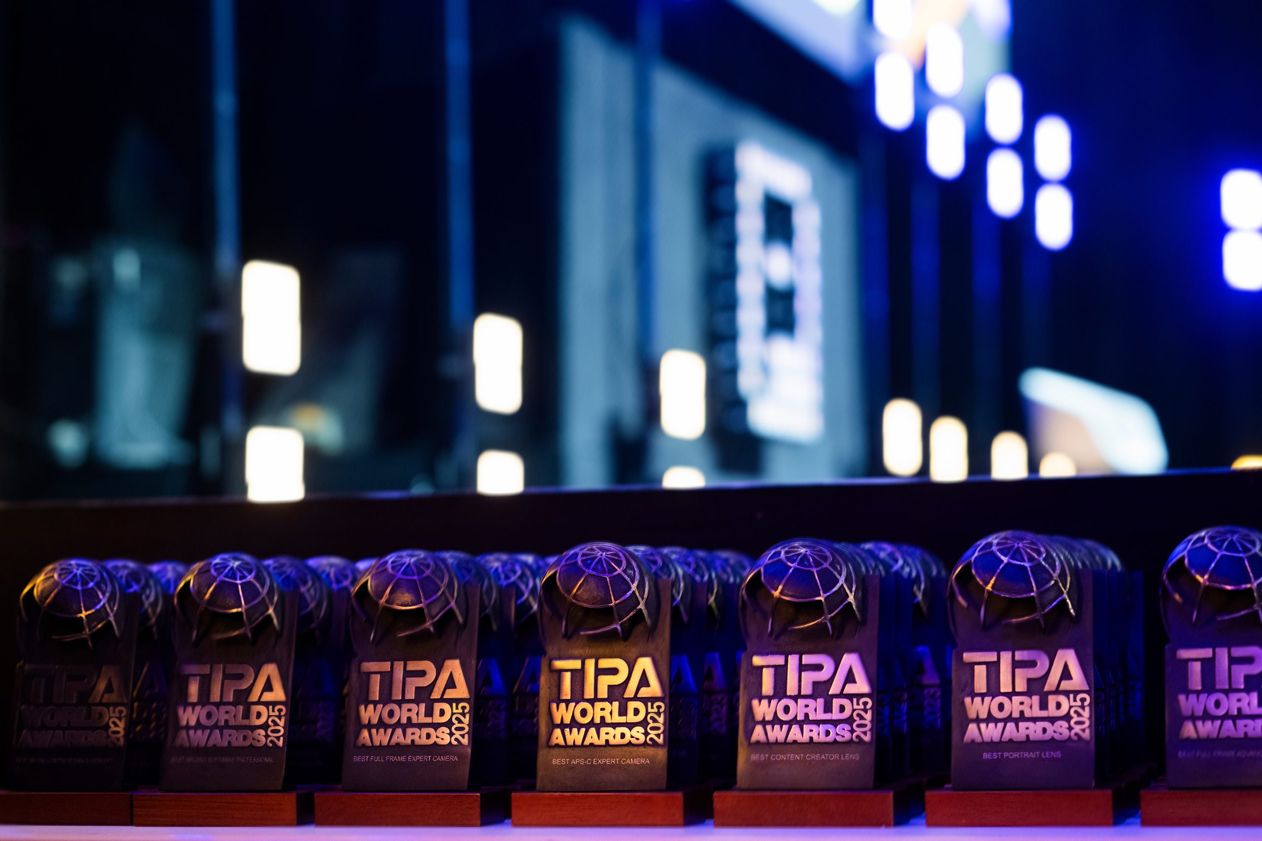 Glittering honors at Imaging World 2025: TIPA World Awards 2025 in the spotlight A row of gleaming TIPA World Awards 2025 trophies on an illuminated table, with a stylishly lit stage in the background during the awards ceremony at Imaging World 2025.