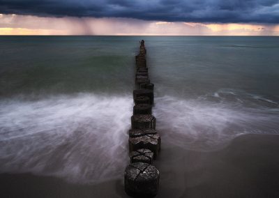 Wellen treffen auf Buhnenreihe, im Hintergrund Regenwand und dramatischer Himmel.
