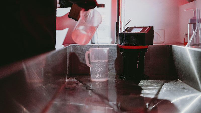 Imaging World Workshop: Color Film Developing – Praxis im Fotolabor Nahaufnahme einer Filmszene im Labor: Hände gießen Flüssigkeit aus einem Messbecher in eine Entwicklungseinheit mit AGO Film Processor, im Hintergrund rotes Sicherheitslicht