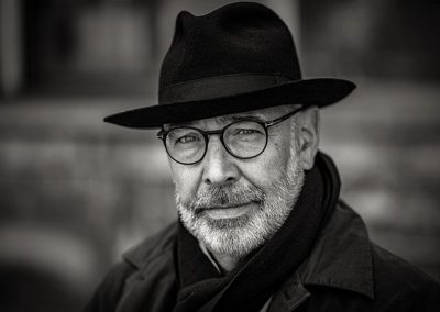 Schwarz-Weiß-Porträt eines Mannes mit Hut und Brille vor unscharfem Hintergrund.