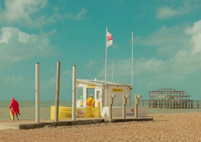 Strandszene mit Rettungsschwimmerhäuschen, zwei Personen am Kiesstrand und den Überresten eines Piers im Hintergrund.