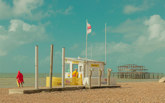 Strandszene mit Rettungsschwimmerhäuschen, zwei Personen am Kiesstrand und den Überresten eines Piers im Hintergrund.
