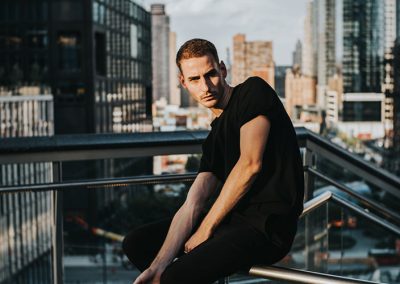 Mann in schwarzem Shirt posiert auf Dachterrasse mit Skyline im Hintergrund – modernes urbanes Portrait.
